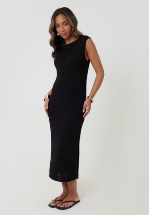 Mujer con un vestido midi de punto negro sin mangas, chanclas negras, joyas de oro, de pie frente a un fondo claro y liso.