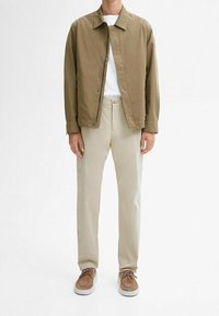Uomo vestito con una giacca casual marrone, una maglietta bianca, pantaloni beige e scarpe in suede marrone, in piedi contro uno sfondo bianco.