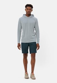 Maglietta a maniche lunghe grigia chiara con cappuccio abbinata a pantaloni corti blu scuro. Il modello indossa sneakers beige con accenti rossi. Design semplice e informale.