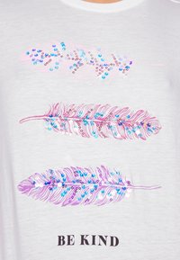 Camiseta blanca con tres gráficos de plumas en rosa y púrpura con detalles en azul, adornada con lentejuelas. El texto dice "SÉ AMABLE" debajo.