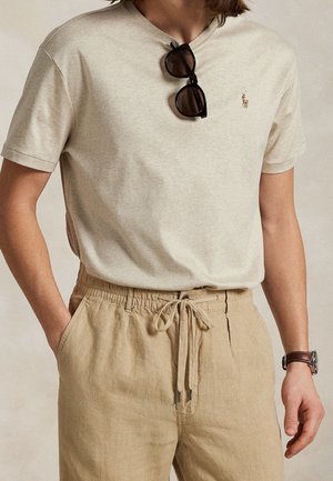 Personne portant une chemise beige à manches courtes avec des lunettes de soleil accrochées au col, un pantalon beige à cordon et une montre-bracelet marron.