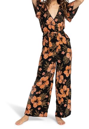 Vrouw draagt een zwarte jumpsuit met brede pijpen en oranje bloemenprint, met een ceintuur in de taille en korte, uitlopende mouwen, staat blootsvoets.