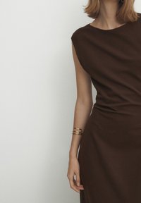 Ruskea hihaton mekko, jossa on laiva-kaula-aukko, tiukka siluetti ja pehmeä tekstuuri. Kultaiset korut korostavat kättä, joka lepäilee vyötäröllä.