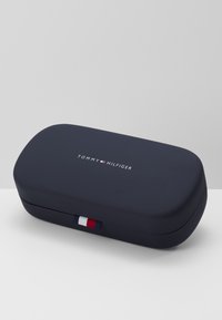 Étui pour lunettes rigide bleu marine avec une texture lisse, arborant un logo blanc et un accent rayé rouge, blanc et bleu sur le côté.