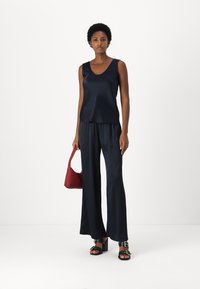 Navy Satin Ärmelloses Top mit Rundhalsausschnitt, kombiniert mit lockeren navyblauen Hosen. Das Model hält eine rote Tasche und trägt schwarze, hochhackige Sandalen.