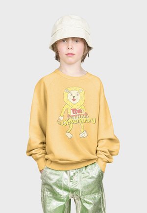 Geel sweatshirt met een leeuwgrafiek, "The Animals Observatory" tekst in het rood, gecombineerd met glanzende groene metallic broek en een witte bucket hat.