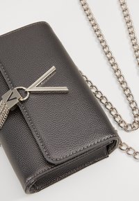 Šedá texturovaná peněženka s kovovým řetězovým popruhem, na předním klopě zdobená stříbrným klenotem ve tvaru písmene V.