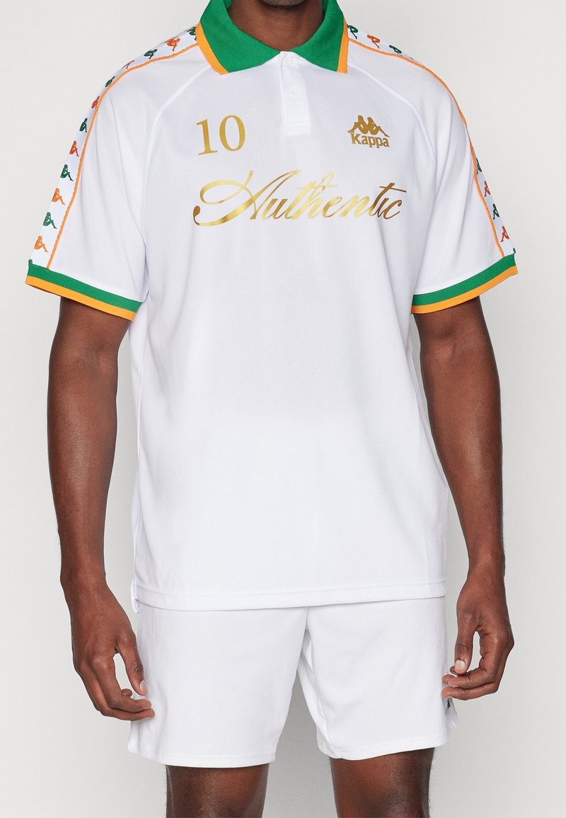 Bílá polo košile se zeleným límcem, oranžovými akcenty a zlatým nápisem. Obsahuje logo, číslo 10 a vzorované rukávy. Spárováno s bílými šortkami.