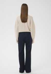 Femme aux longs cheveux bruns et raides, portant un pull beige et un pantalon large bleu marine, debout de dos sur un fond blanc uni.
