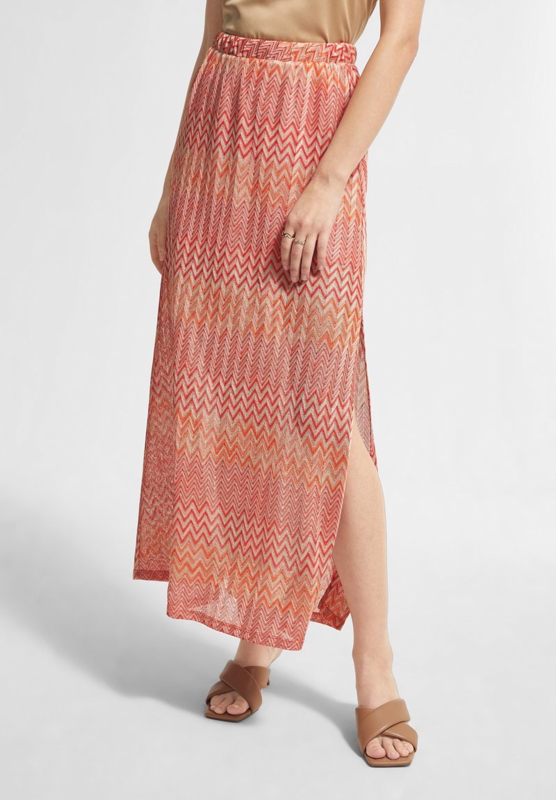 Jupe à motif zigzag diagonal long, rouge et orange, en tissu transparent, avec taille élastique et fente sur le côté, portée avec des sandales plates marron.