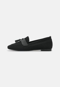Zapato negro de malla tipo slip-on con suela plana, parte superior texturizada y una correa decorativa con flecos. Diseño minimalista con un acabado suave.
