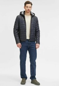 Giacca trapuntata grigio scuro con cappuccio, dotata di zip e polsini elasticizzati, abbinata a una maglietta chiara e jeans blu. Sneakers verde oliva.