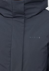 Veste matelassée gris foncé avec un col haut et un logo MAZINE visible. Texture lisse avec un design élégant et moderne et une silhouette épurée.