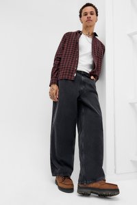 Mlad moški se opira ob belo steno, oblečen v rdečo karirano srajco, belo majico brez rokavov, široke črne hlače, rjave čevlje in srebrno verižico.