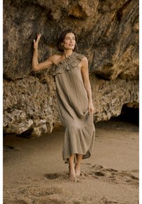 Olive roheline maxi kleit, millel on üks volangiga õlg, pleeritud kangas ja laienev alläär. Jalas paljajalu liivase ranna ääres kalju vastu.