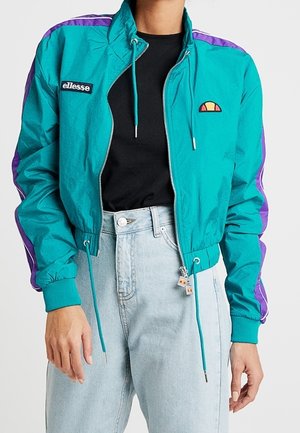 Aquafarvet og lilla jakke med lynlås og ellesse-logoer over en sort skjorte, kombineret med lyseblå højttaljede jeans.