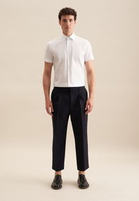 Witte korte mouwen overhemd met knoopjes, gecombineerd met hooggewaisted marineblauwe geplooide broek en zwarte veterschoenen, gepresenteerd tegen een neutrale achtergrond.
