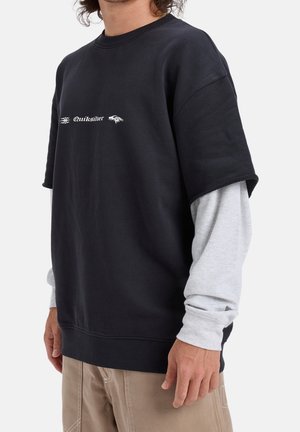 Person trägt mehrlagige Kleidung mit einem schwarzen Quiksilver-Kurzarm-Sweatshirt über einem hellgrauen Langarmshirt und beigen Hosen.