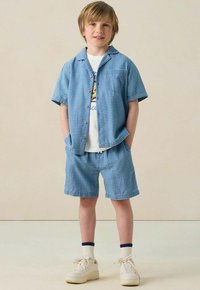 Junge trägt ein blau gestreiftes Kurzarmhemd und passende Shorts mit einem weißen Grafik-T-Shirt, Socken und beigen Turnschuhen, steht mit den Händen in den Taschen.