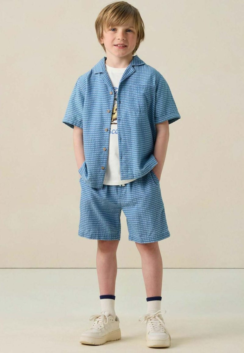 Junge trägt ein blau gestreiftes Kurzarmhemd und passende Shorts mit einem weißen Grafik-T-Shirt, Socken und beigen Turnschuhen, steht mit den Händen in den Taschen.