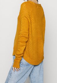 Kvinna som bär en senapsgul stickad tröja och ljusblå jeans, sedd bakifrån mot en enfärgad bakgrund.