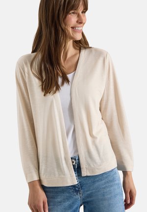 Femme souriante portant un cardigan ouvert beige clair sur un haut blanc et un jean bleu sur un fond uni.