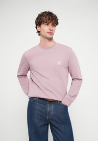 Svetlofialová mikina s dlhými rukávmi, okrúhlym výstrihom a malým štvorcovým logom na hrudi. Kombinovaná s modrými rifľovými nohavicami.