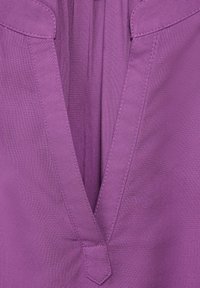 Tessuto viola con un profondo scollo a V e arricciature sul petto. Texture liscia, bordi rifiniti e sottili dettagli di cucitura.