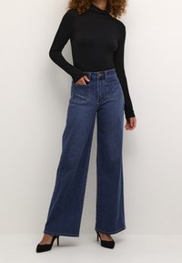 Jeans in denim a gamba larga di colore blu scuro, con tasche anteriori e posteriori, vita alta e cuciture a contrasto, abbinati a un top nero a maniche lunghe.