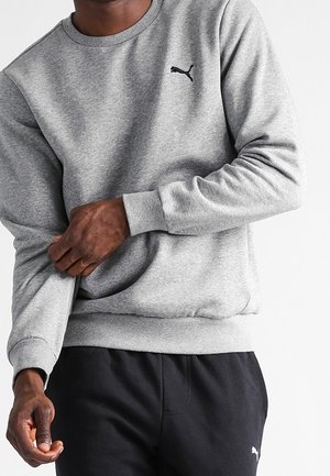 Mężczyzna poprawiający rękaw szarej bluzy Puma, ubrany w czarne spodnie Puma, na jednolitym białym tle.