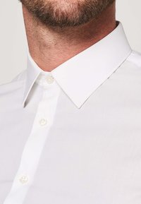 Camicia bianca con colletto classico, texture liscia e bottoni simili a perle. Non ci sono motivi o accenti visibili.