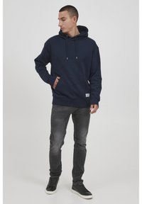 Navy Kapuzenpullover mit Kängurutasche, Zugkordel und gerippten Bündchen. Kombiniert mit grauen Jeans und schwarzen Turnschuhen.