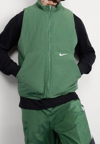 Person med en grön zip-up väst med fickor över en svart långärmad skjorta, tillsammans med gröna byxor som har ett vitt sidostreck.