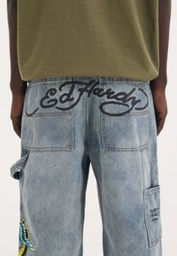 Ljust blå denim cargobyxor med svart "Ed Hardy" text på den bakre midjebandet. Har en sidoficka med texten "Don't Hate" och färgglad broderi.