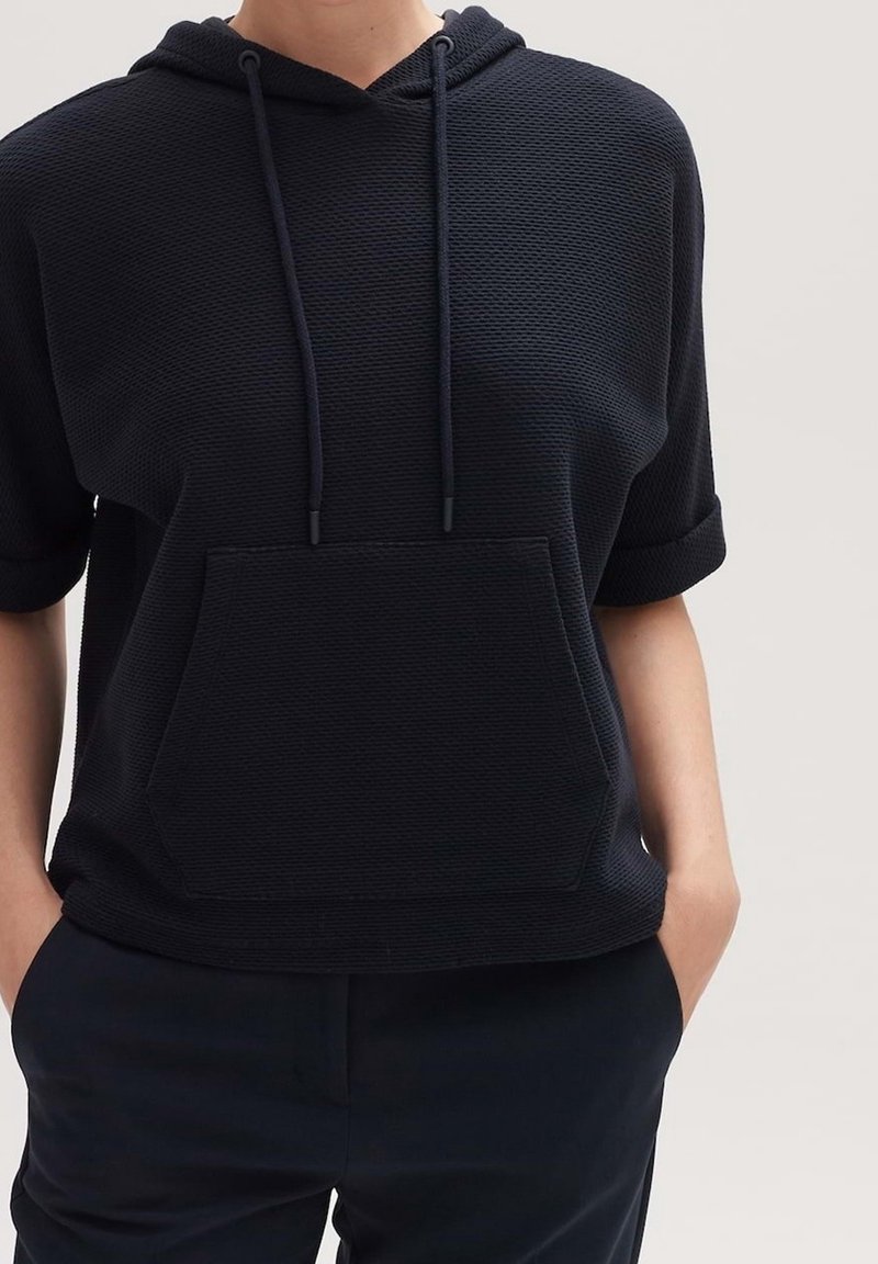 Personne portant un sweat à capuche texturé noir à manches courtes avec poche avant et cordons, mains dans les poches d’un pantalon foncé sur un fond uni.