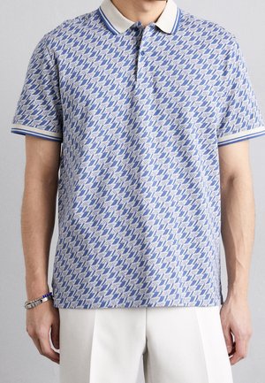 Mand iført kortærmet blå og hvidt mønstret poloshirt med krave og hvide bukser, stående mod en grå baggrund.