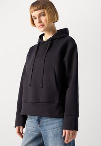 Felpa nera con cappuccio e tasca frontale, caratterizzata da una vestibilità comoda, polsini a costine e una texture liscia. Abbinata a jeans azzurri chiari.