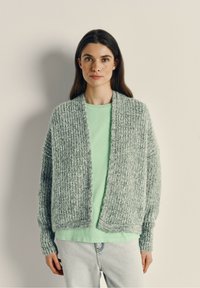 Lichtgrijze, chunky gebreide cardigan met een open voorkant, geribde manchetten en een gestructureerde stof, gedragen over een mintgroene top en lichte denim jeans.