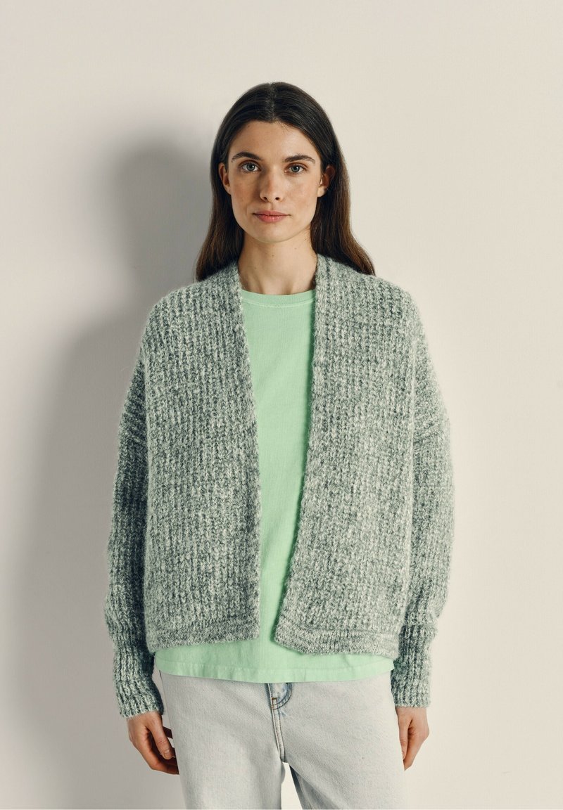 Lichtgrijze, chunky gebreide cardigan met een open voorkant, geribde manchetten en een gestructureerde stof, gedragen over een mintgroene top en lichte denim jeans.