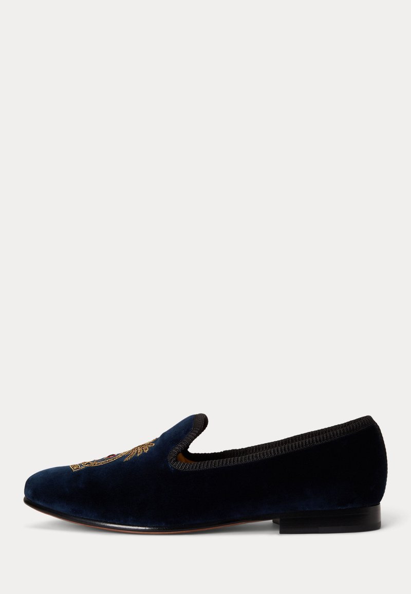 Mocassin en velours bleu foncé avec une bordure noire, un petit talon et un blason brodé en or sur le dessus, présenté de profil sur fond blanc.