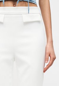 Κοντινή λήψη ενός ατόμου που φοράει ψηλόμεσα λευκά παντελόνια με τσέπες με καπάκι και ένα κοντό τζιν μπουφάν, με ορατό τον κορμό και το δεξί χέρι.