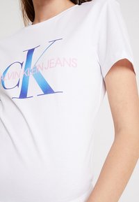 Biała bawełniana koszulka z dużym gradientowym logo "CK" w niebieskim i różowym kolorze, z napisem "CALVIN KLEIN JEANS" poniżej w mniejszej czcionce.