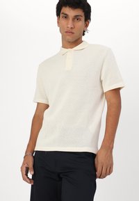 Short-sleeve polo shirt in cream with a textured mesh design and a contrasting light-colored collar. Three-button placket, relaxed fit.