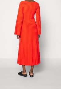 Robe midi rouge vif côtelée à manches longues et col rond. Accompagnée de chaussures noires sans lacets. Tissu texturé avec des rayures verticales.