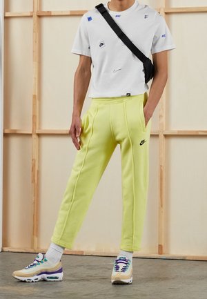 Osoba ubrana w białą koszulkę z logo Nike, żółte spodnie dresowe Nike, wielokolorowe sneakersy i czarną torbę na ramię, stojąca w pomieszczeniu.