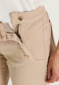Pantalones de algodón beige con una superficie texturizada, que cuentan con un cierre lateral y botones en tono dorado. Primer plano de la cintura y detalle del bolsillo.