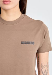 Lyhythihainen ruskea puuvillapaita, jossa on musta "DICKIES"-logo vasemmassa rinnassa. Sujuva tekstuuri, pyöreä pääntie ja normaali istuvuus.