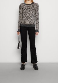 Schwarzer und beiger Pullover mit Muster und Puffärmeln, kombiniert mit schwarzen, ausgestellten Hosen mit Knopfakzenten und klobigen schwarzen Schuhen.