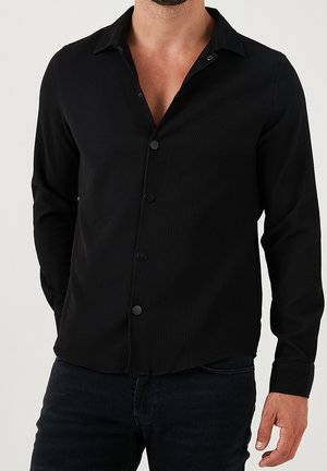 Uomo che indossa una camicia nera a maniche lunghe con bottoni, con una sottile texture a coste e jeans scuri su uno sfondo semplice.
