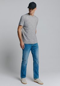 Grijze T-shirt met korte mouwen en een gestructureerde afwerking, gecombineerd met lichtblauwe jeans en witte sneakers. Het model draagt een zwarte pet.